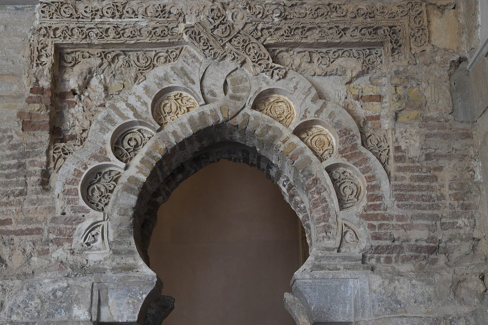 A horseshoe-shaped arch in a brick wall, framed by a scalloped band of carved roundels and panels of interlaced plant scrollwork in plaster.