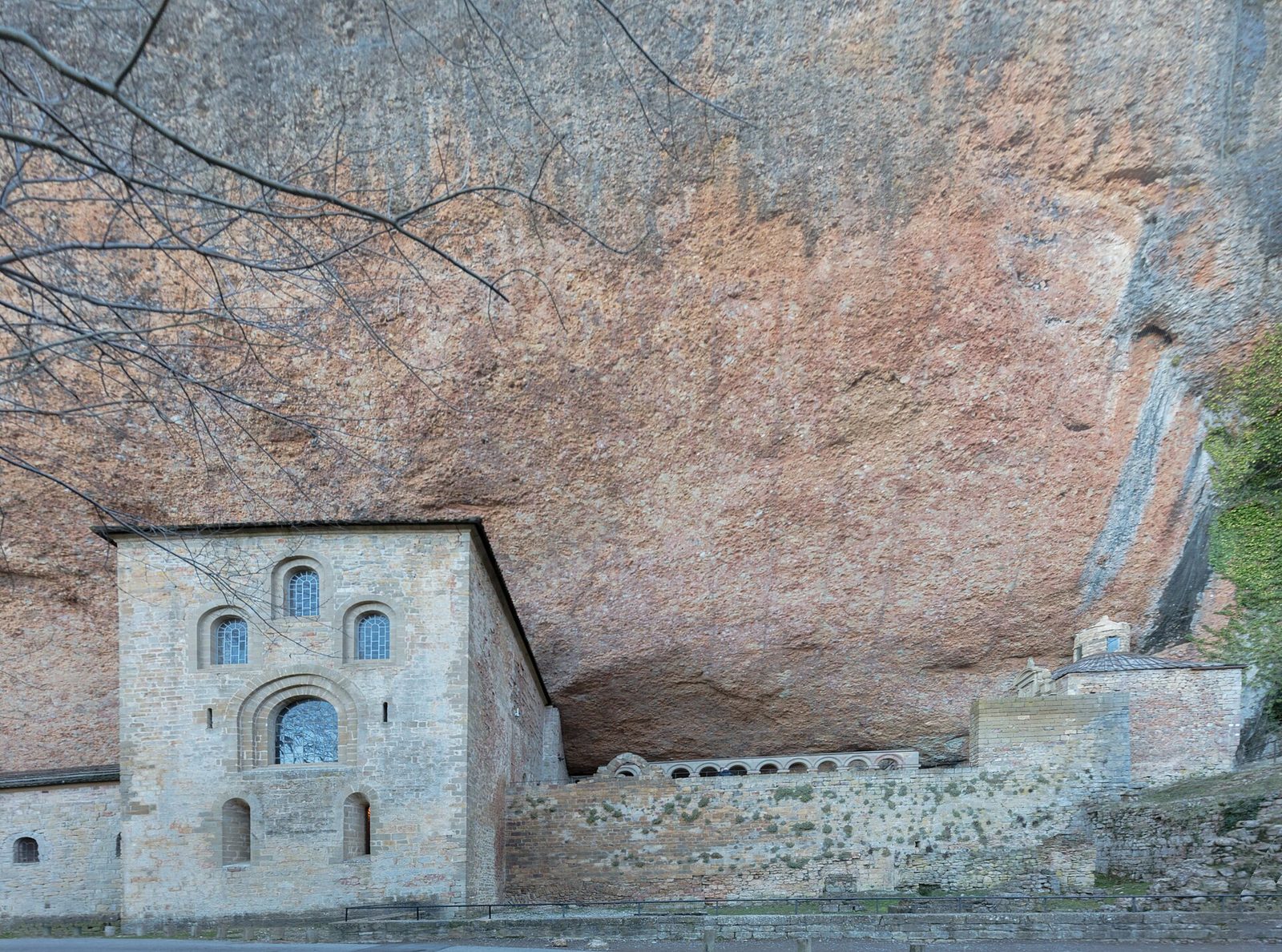 An old stone monastery built tight under a vast overhanging cliff of red rock.