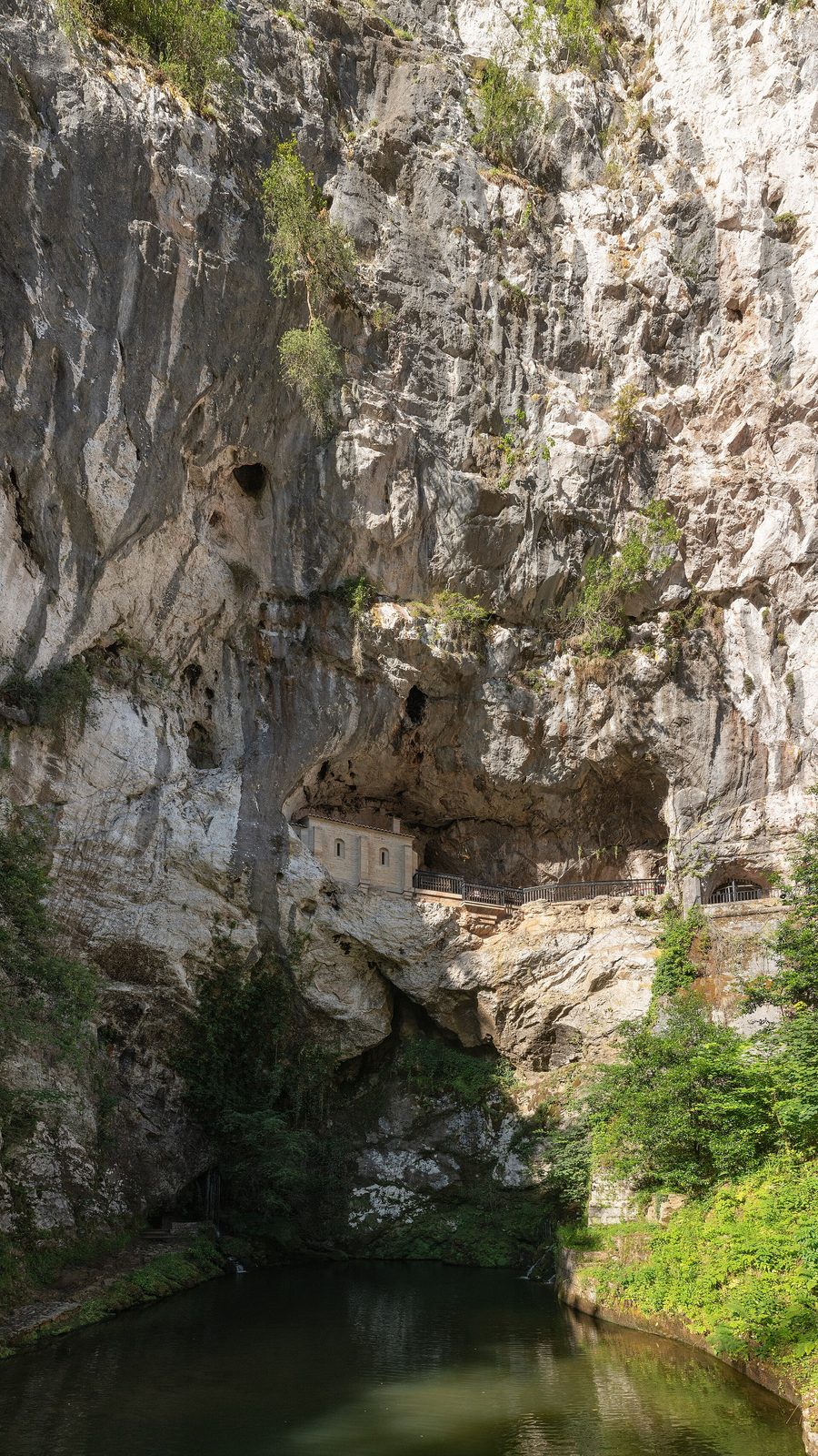 A cave mouth high in a sheer limestone cliff with a small chapel built into it, trees below and a pool at the foot of the rock.