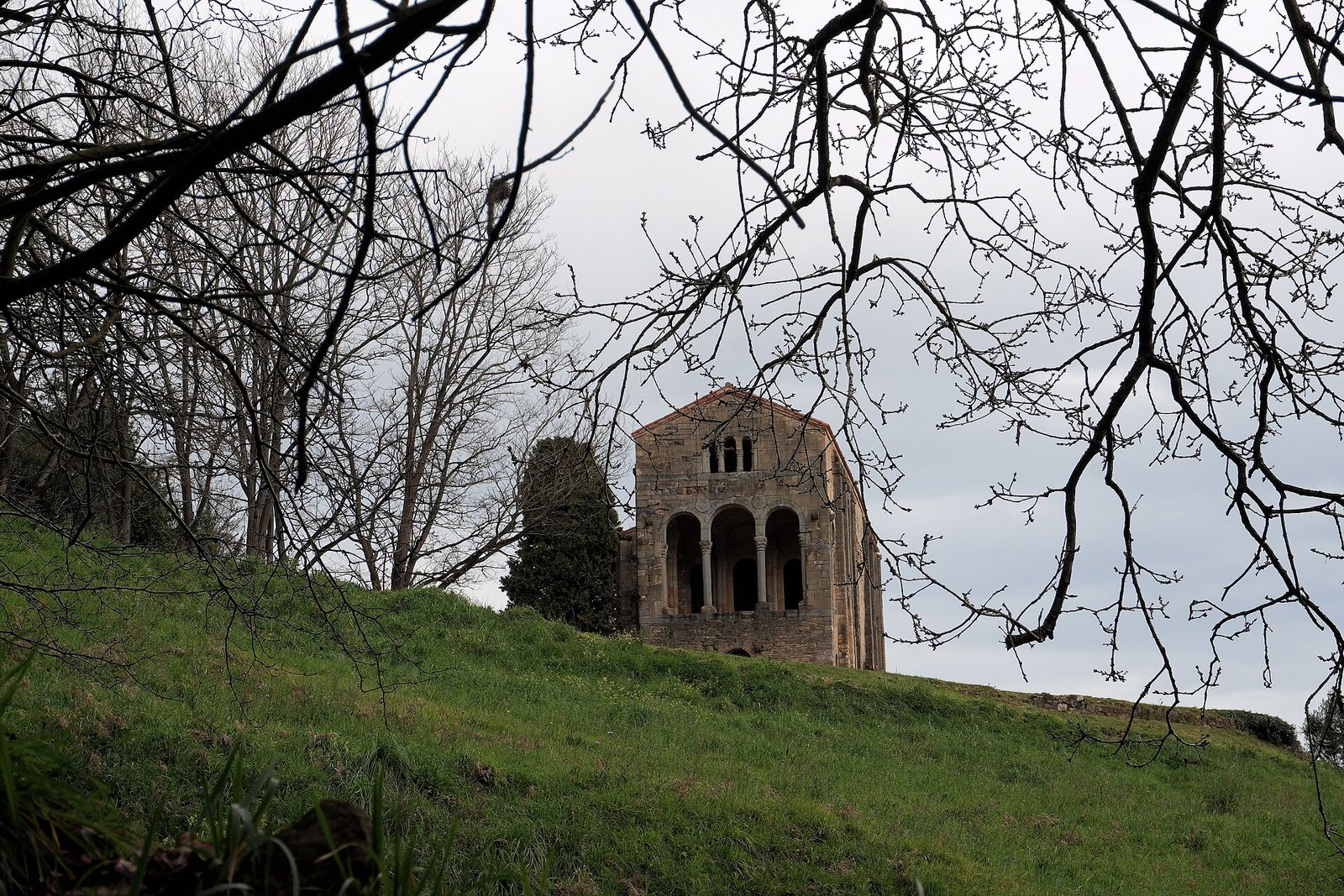 A small rectangular stone hall on a grassy hillside, with buttressed walls and arched windows, seen through bare branches.