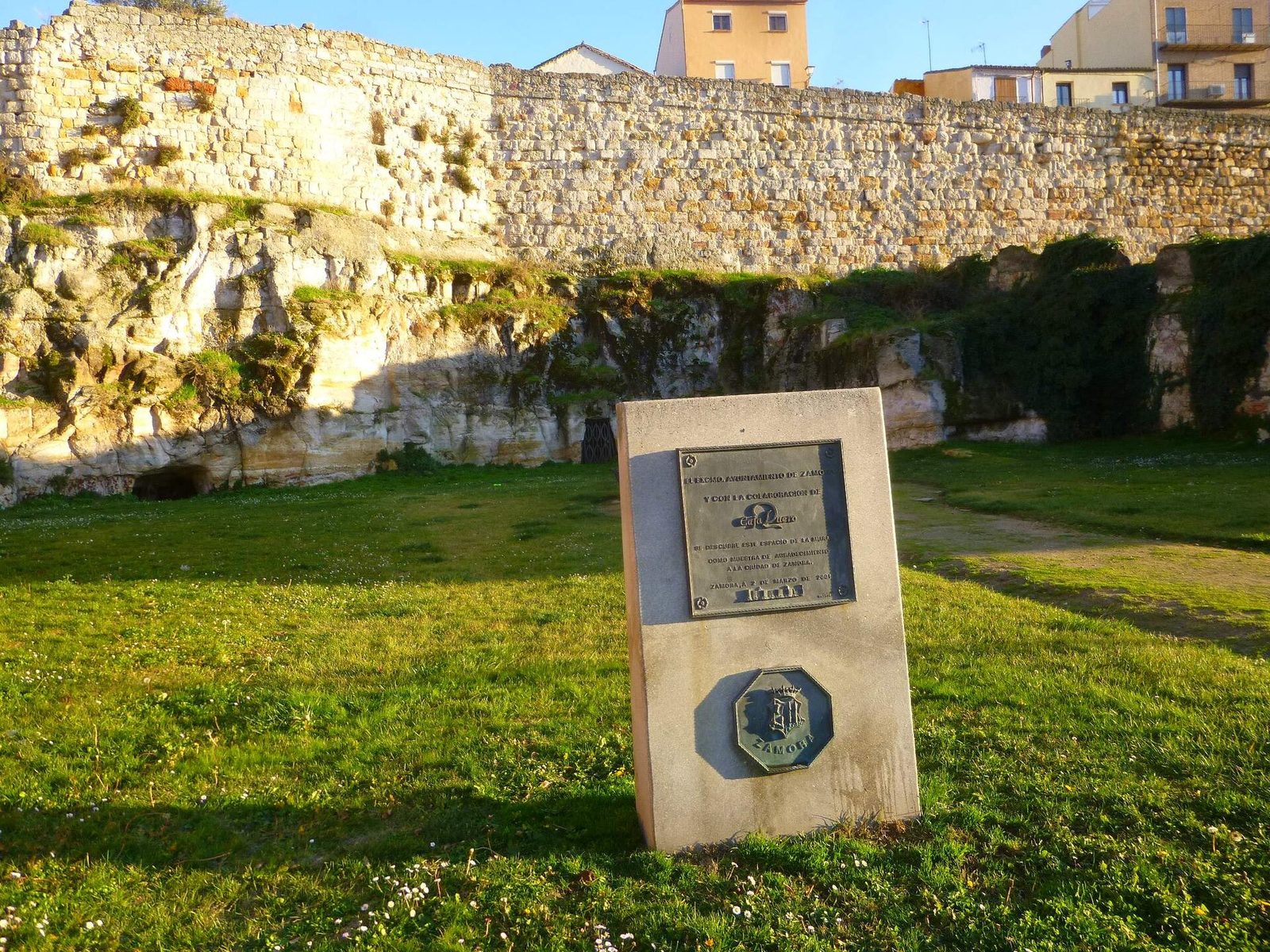 A long stretch of rough stone town wall along a grassy bank, an information panel standing in front of it and buildings above.