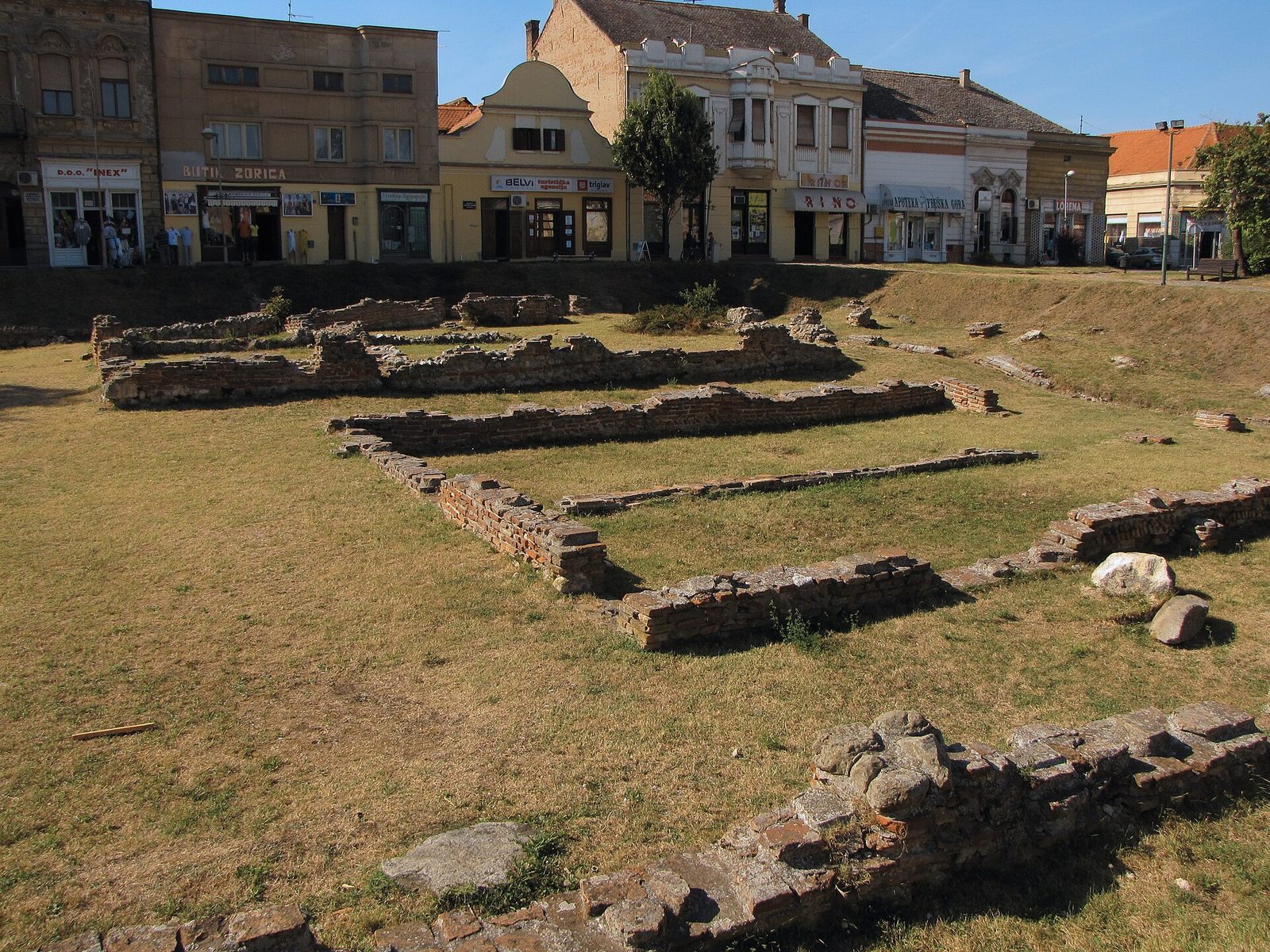 Low brick foundations of Roman walls in a grassy open plot, town houses behind.