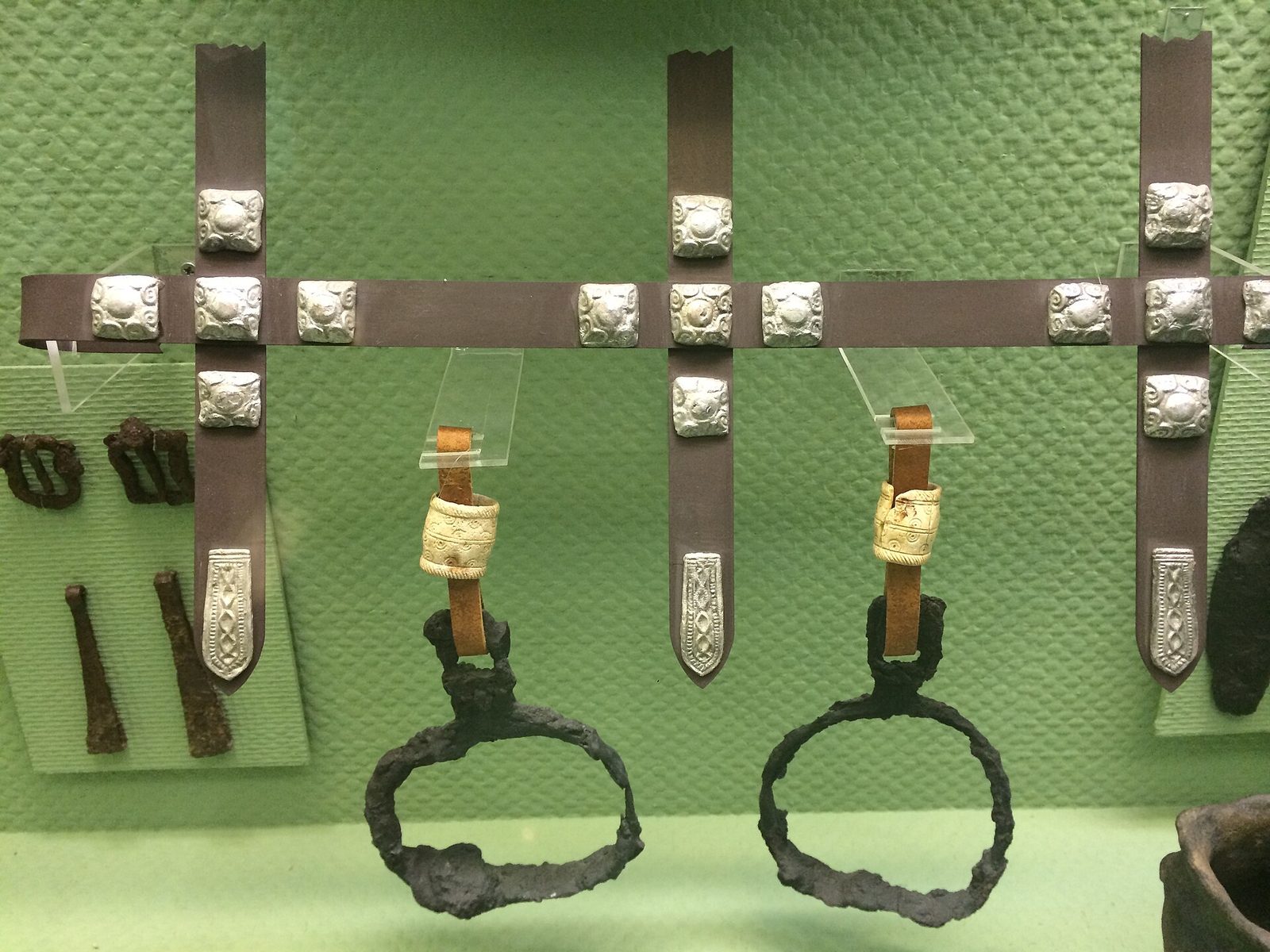 A pair of iron stirrups and leather straps with square silver fittings, mounted on a green museum board.
