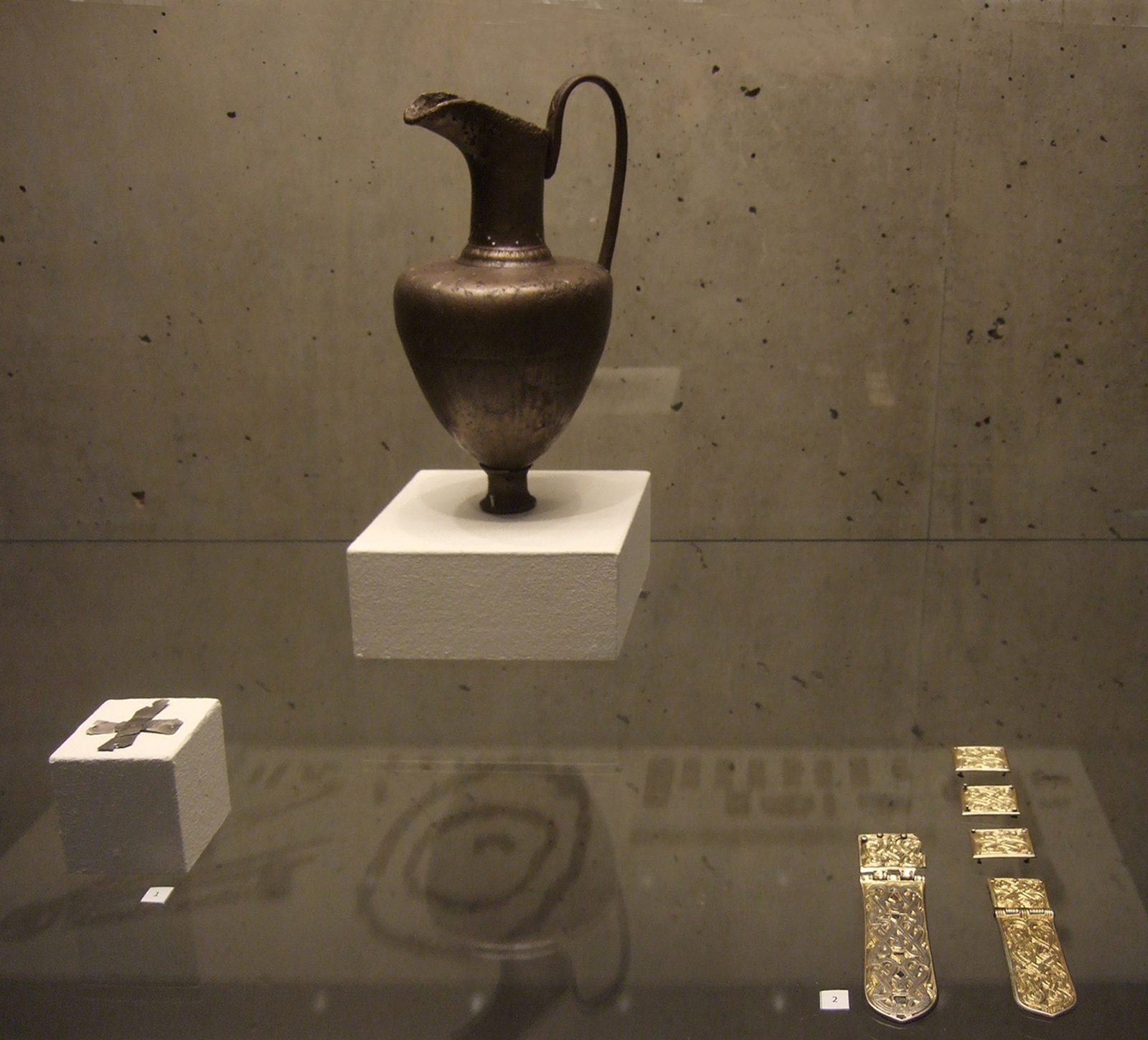 A dark metal jug on a white plinth, with gilt strap ends and a small cross laid out beside it.