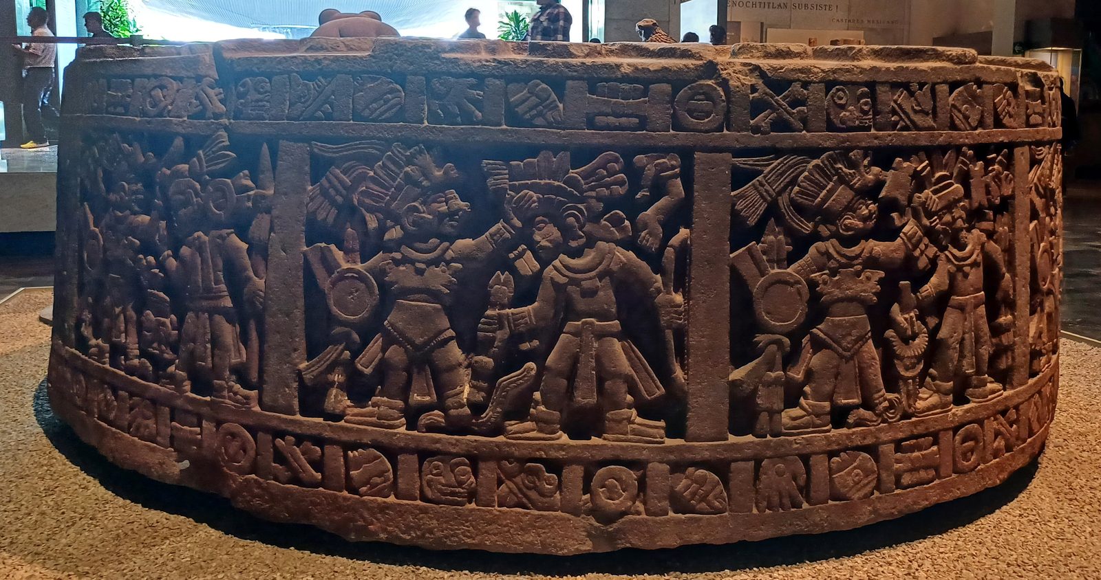 A massive round carved stone with reliefs of paired warriors around its side.