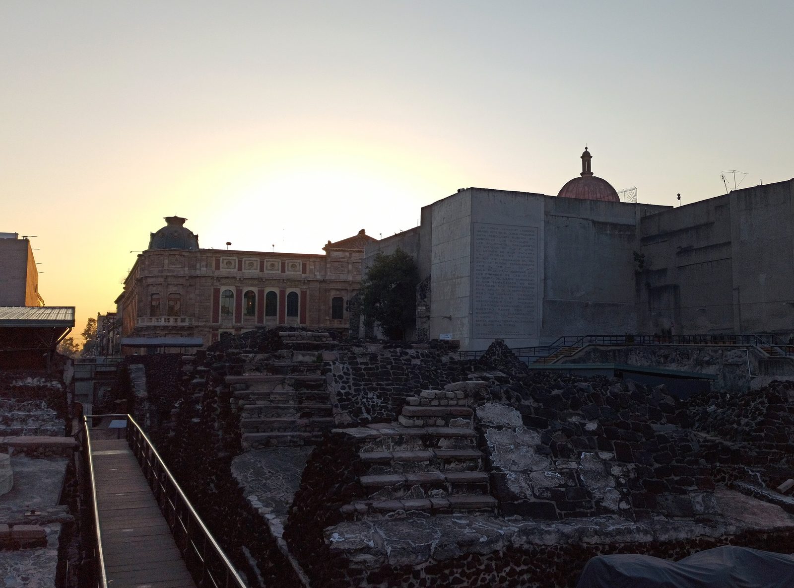Excavated layers of stone stairs and platforms in a city centre at dusk, colonial buildings behind.