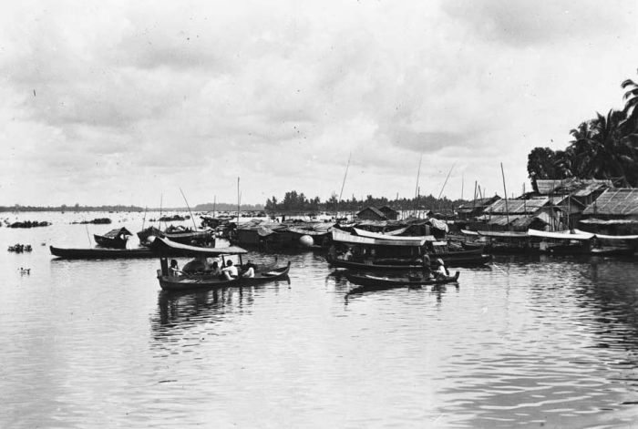 An old photograph of a wide river crowded with small boats and moored praus, houses on piles along the bank.