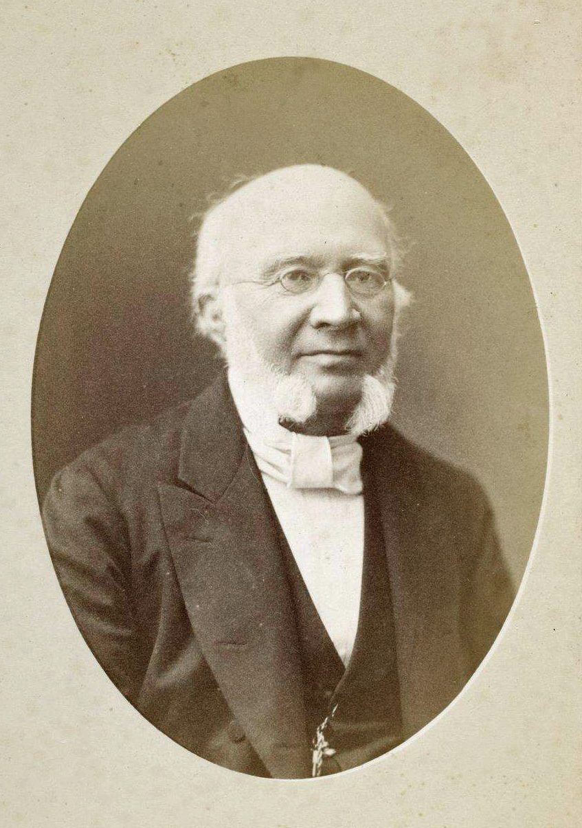 An oval sepia photograph of an elderly man with white side-whiskers and spectacles, in a dark coat.