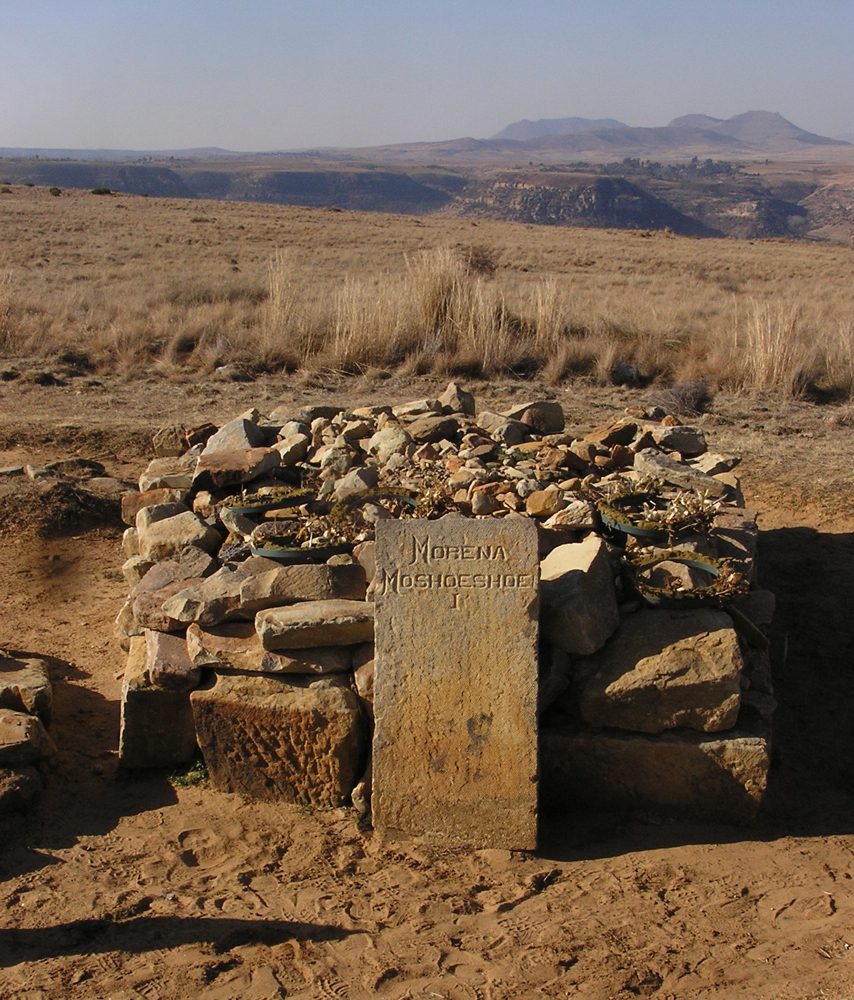 A low cairn of piled stones with an inscribed headstone, on a grassy summit looking out over distant hills.