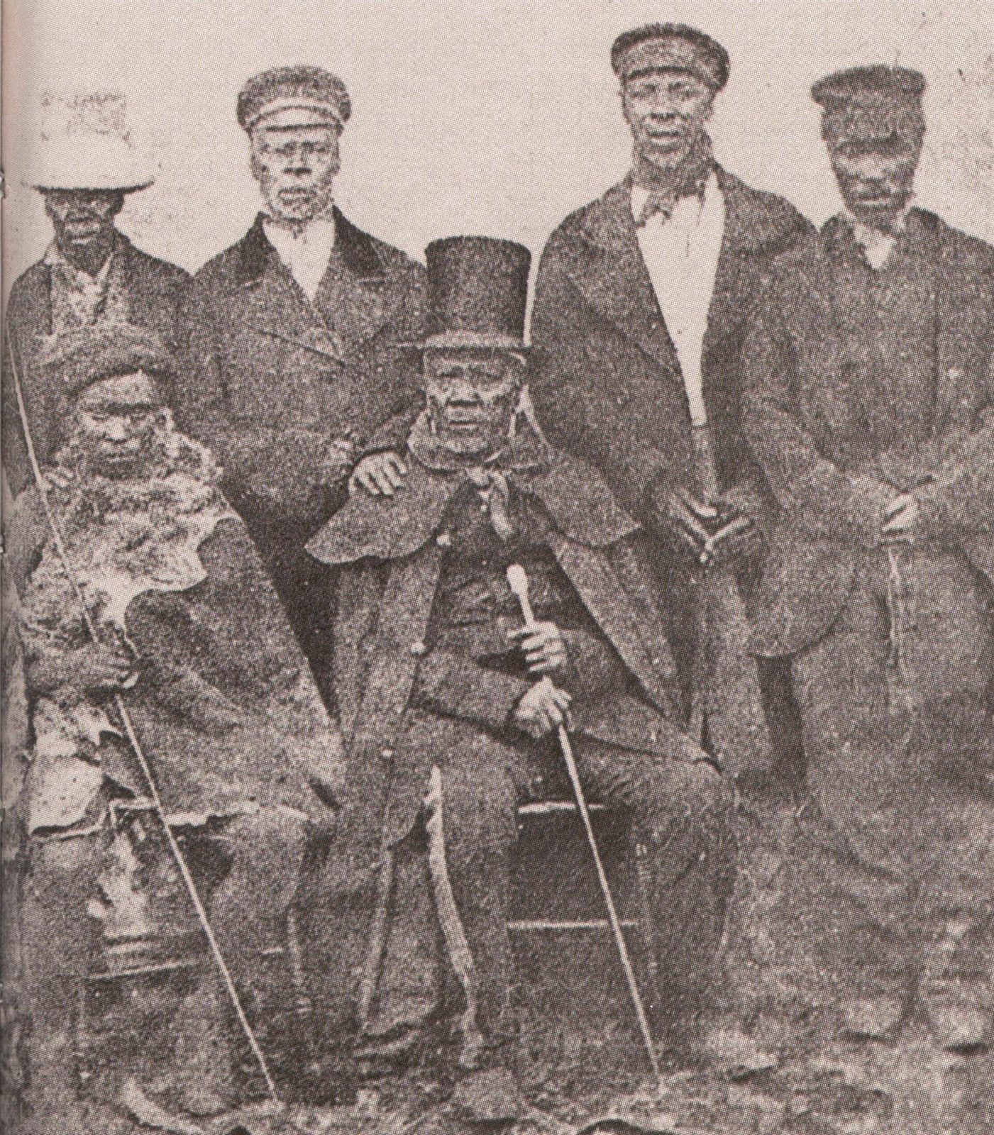 A grainy photograph of six men in coats and caps, two seated, one of them an old man in a top hat holding a cane.