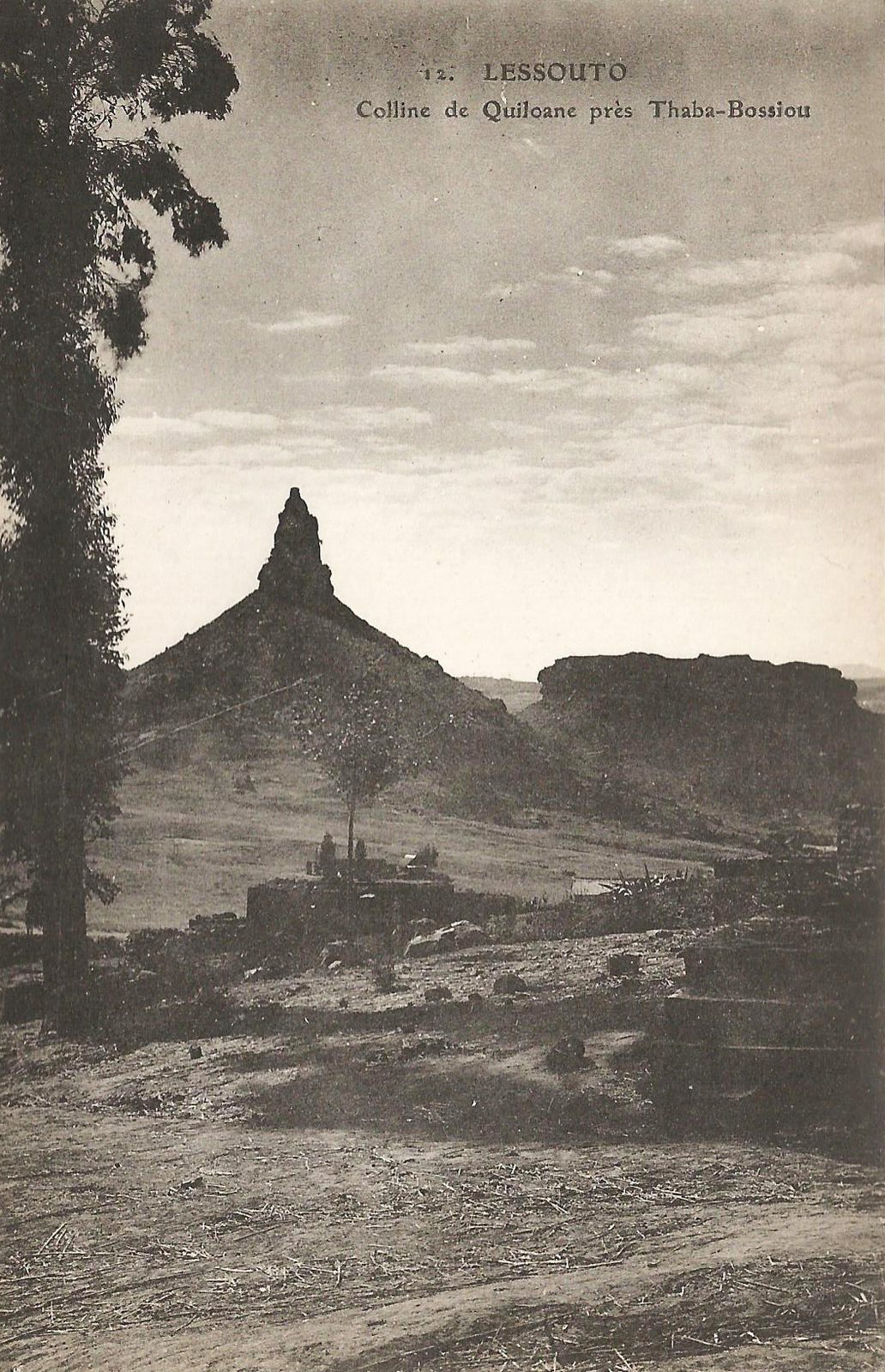 An old photograph of a steep rock pinnacle rising from a plain, a flat-topped cliff behind it.