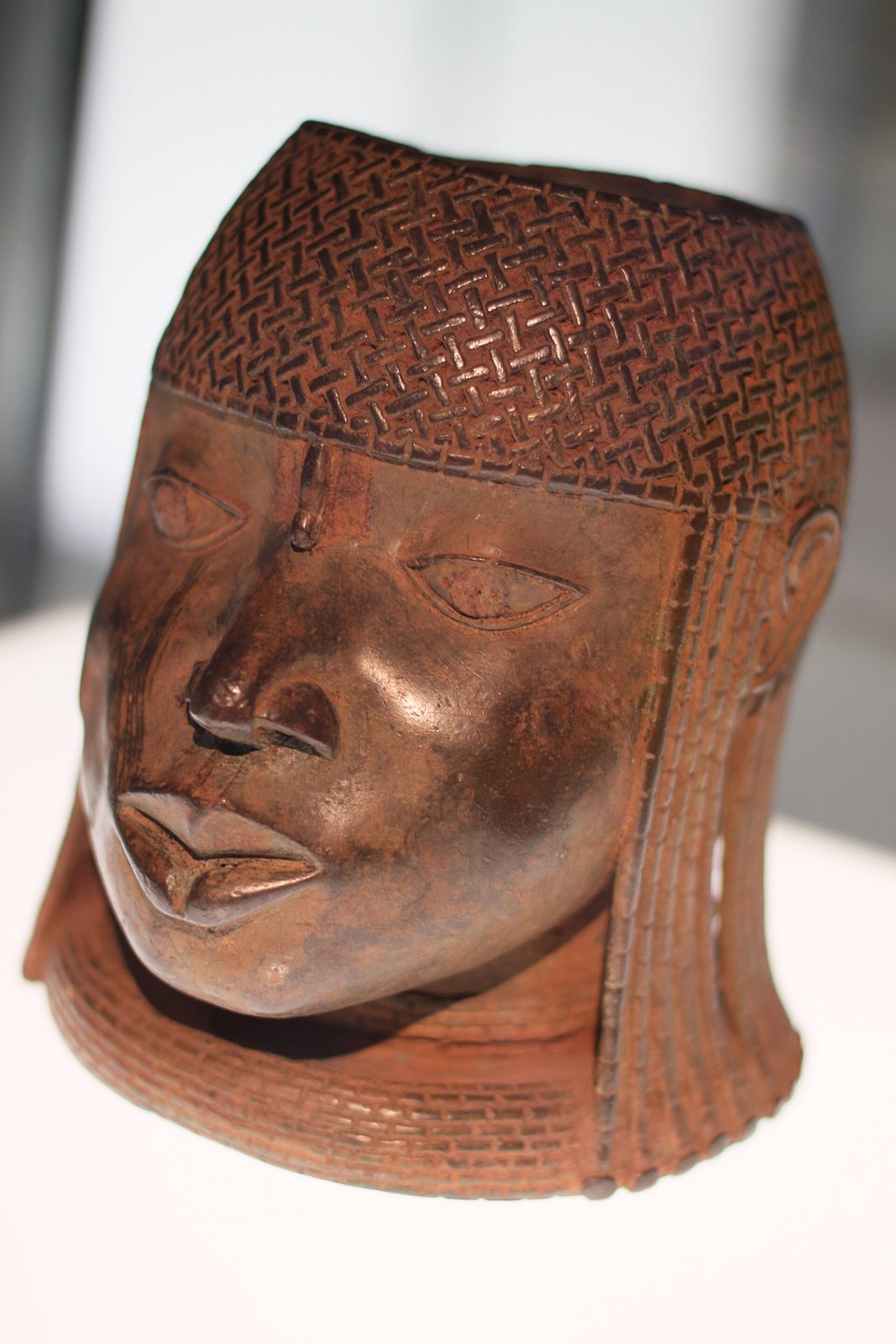 A brass head of a king with a crosshatched cap, lit in a dark museum case.