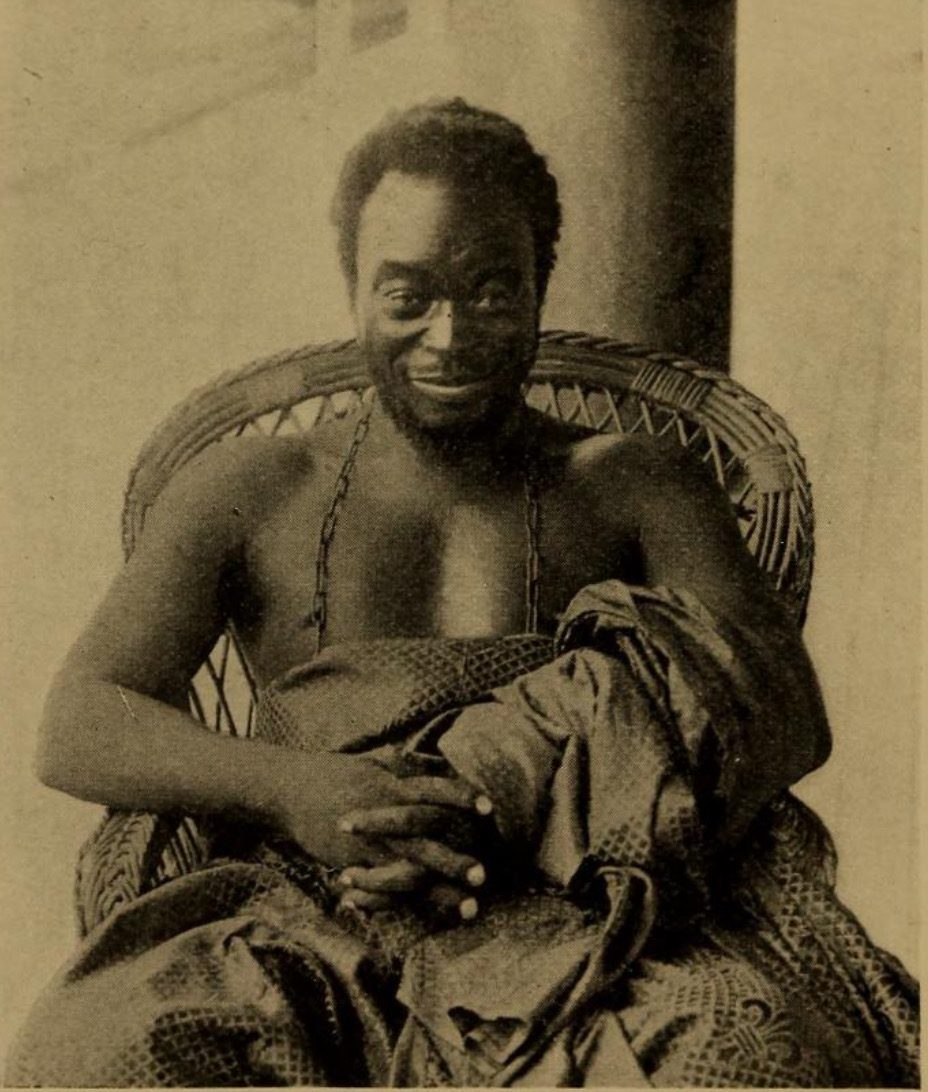 A sepia photograph of a man seated in a wicker chair, wrapped in a patterned cloth, a string of beads at his neck.