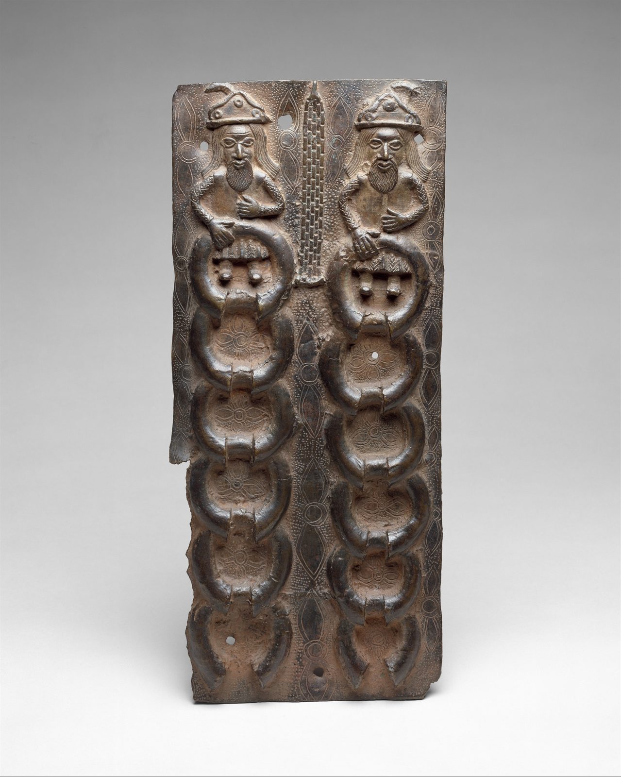 A brass plaque in relief of two small European figures in hats at the top, above two long columns of horseshoe-shaped bracelets.