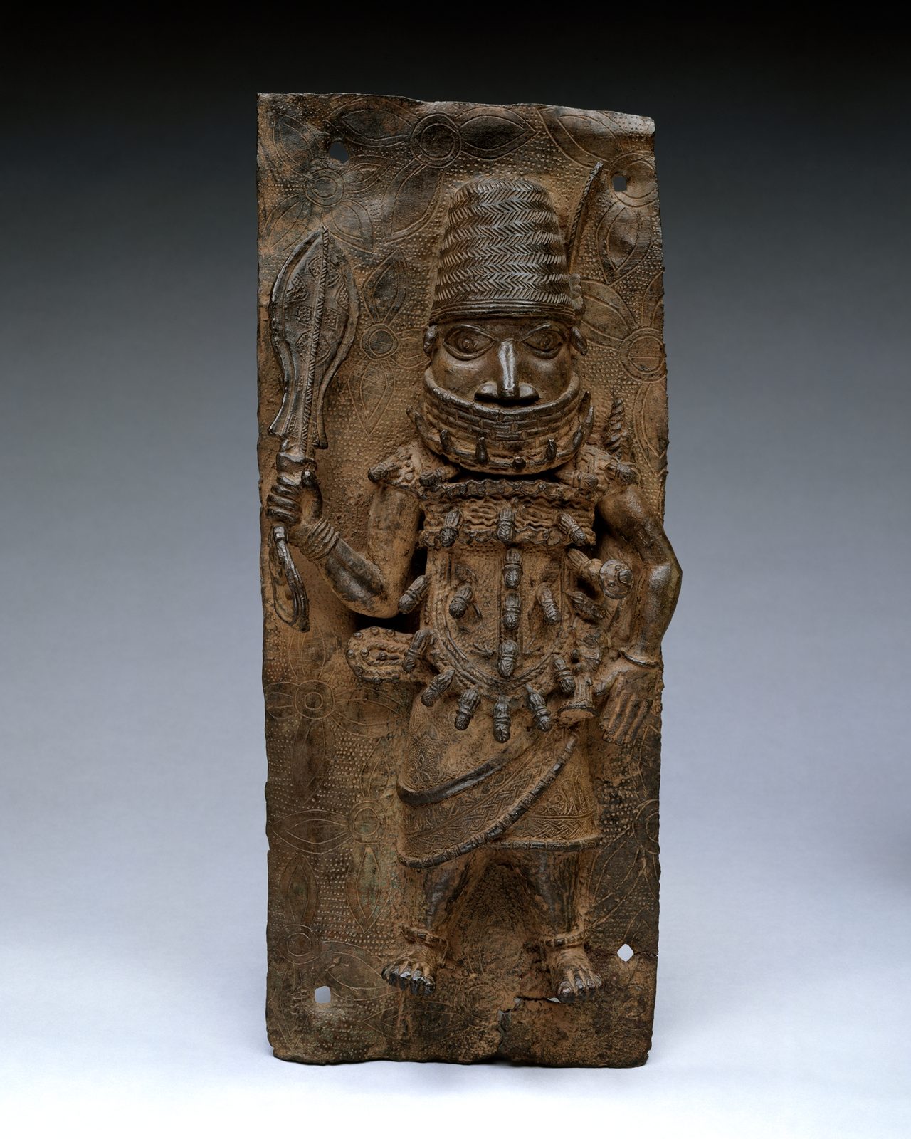 A rectangular brass plaque in relief of a chief standing with a raised sword, wearing a beaded helmet and collar.