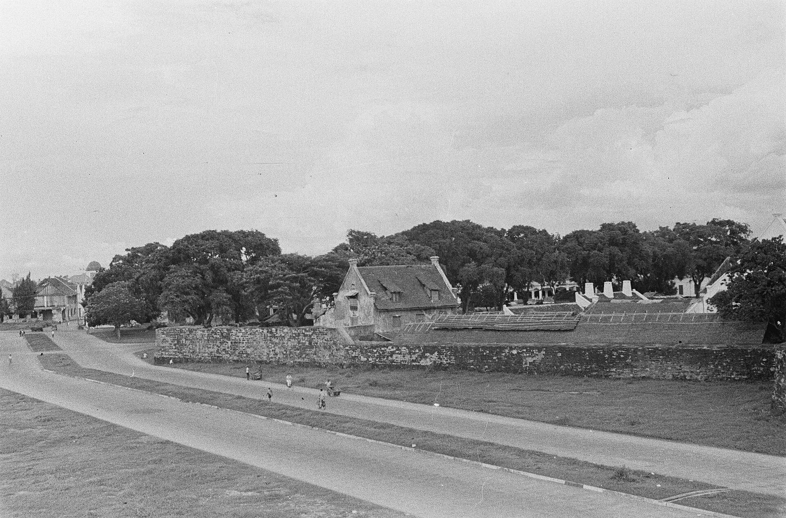 A photograph of a long low fortification wall beside a road, with a gabled building rising inside it and trees beyond.