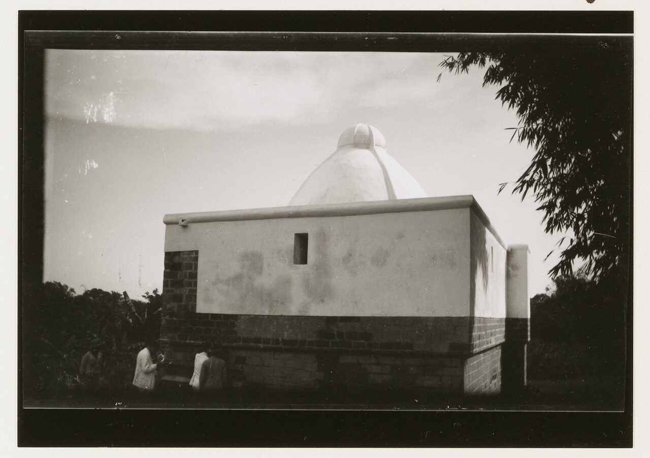 An old photograph of a whitewashed tomb with a domed top standing in a walled enclosure, a figure beside it.