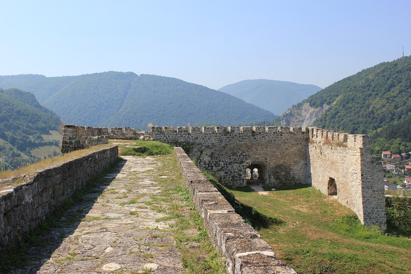 A paved wall-walk leading to a crenellated stone gate tower, wooded hills beyond.