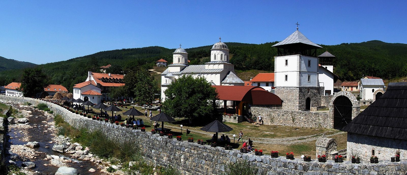 A monastery church and white bell tower beside a stony stream, wooded hills behind.
