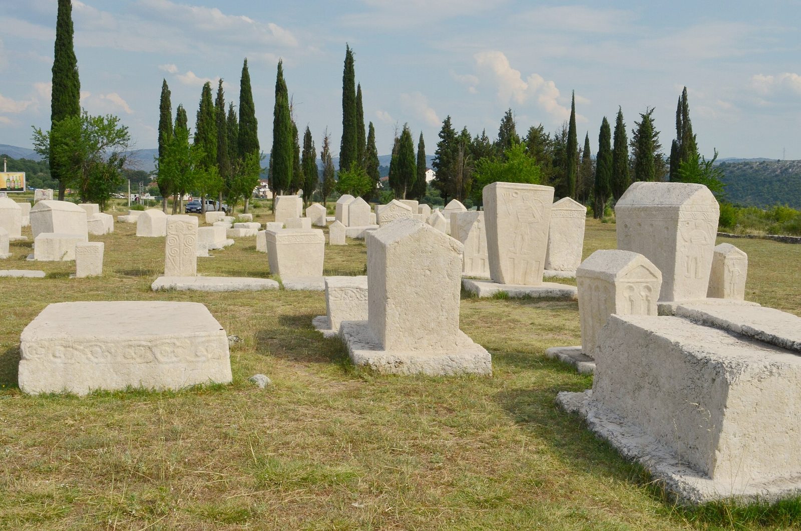 Rows of large carved white tombstones, some shaped like houses, in a meadow lined with cypresses.