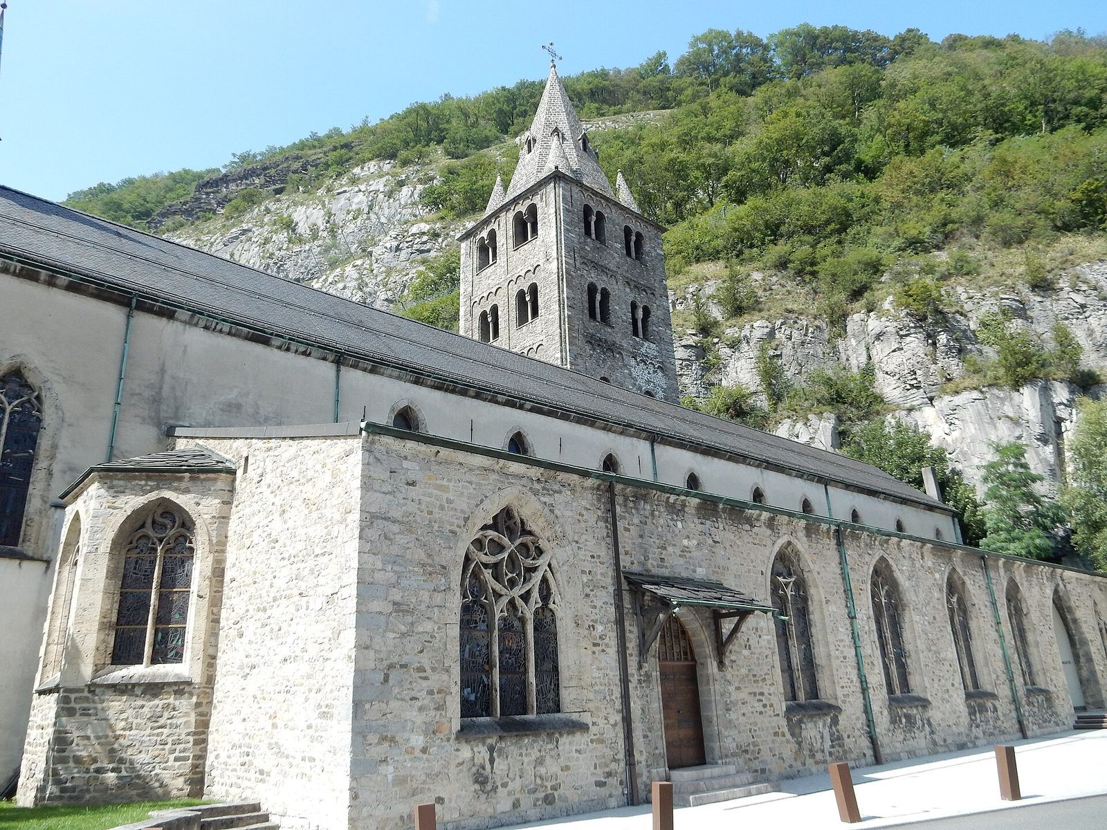 A church with a tall square stone bell tower standing against a sheer wooded cliff that rises directly behind the roofline.
