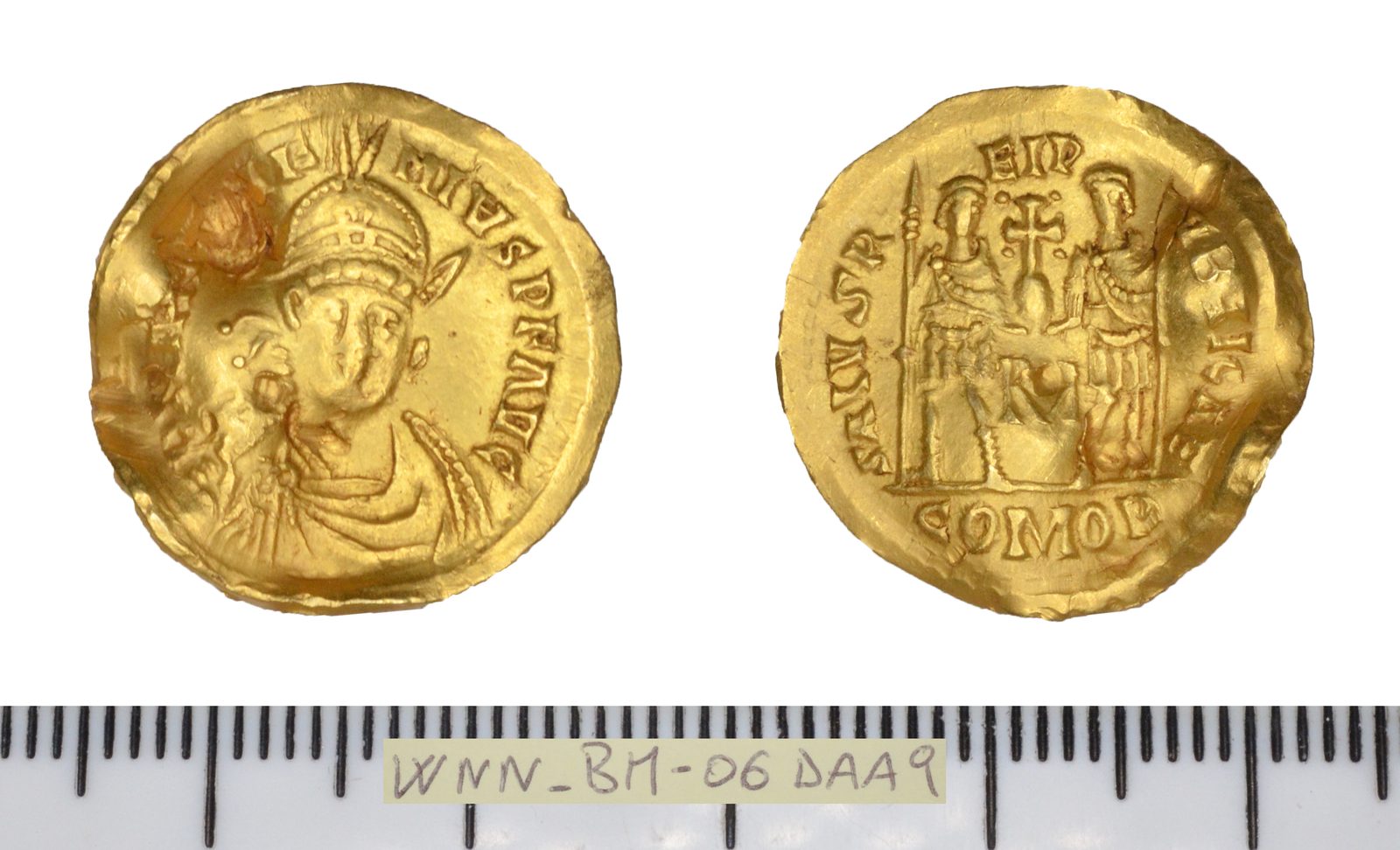 The two faces of a thin gold coin photographed above a ruler: a helmeted bust facing forward holding a spear, and two standing figures supporting a cross between them.