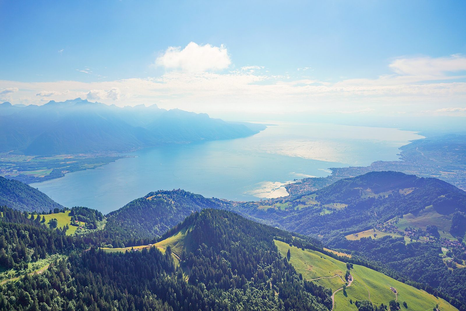 A long lake lying between steep wooded slopes, with snow-covered Alpine peaks rising beyond the far shore under a pale sky.