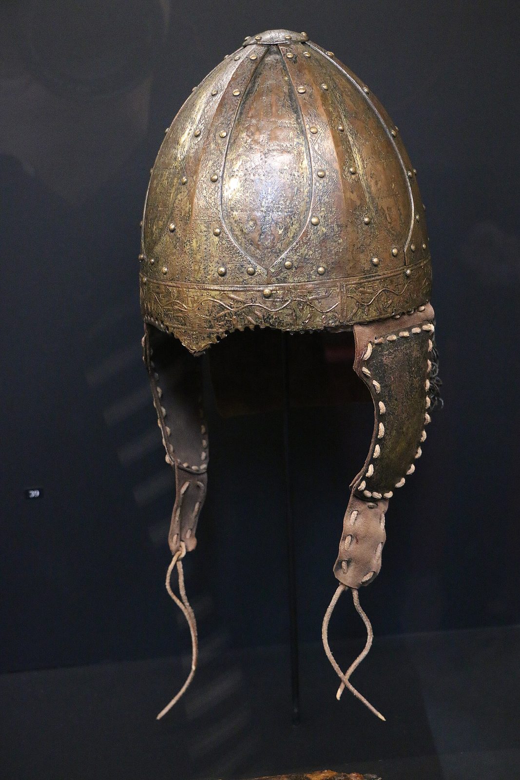 A conical iron helmet built from curved plates riveted to a frame, much corroded and incomplete, mounted on a stand in a museum case.