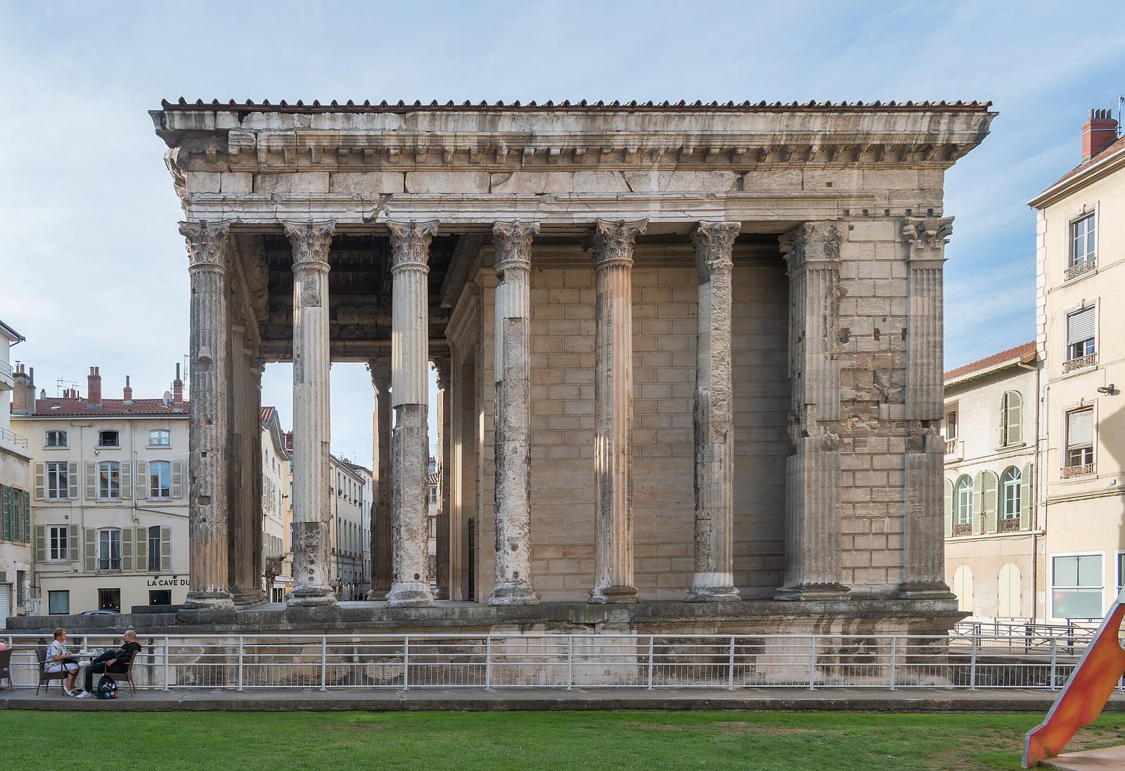 A small Roman temple of Corinthian columns on a high podium, its pediment and entablature intact, standing in a paved square among later town buildings.