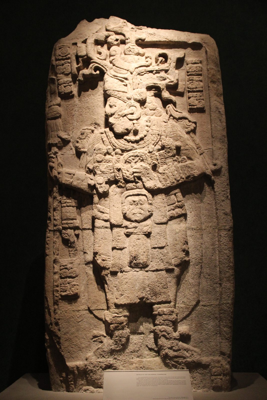 A tall carved stone stela of a richly dressed standing ruler, lit against a dark background.