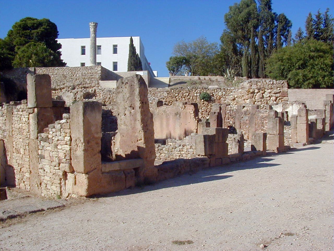 Ruined walls of dressed stone blocks and rubble lining a gravel path, a white modern building and trees behind.