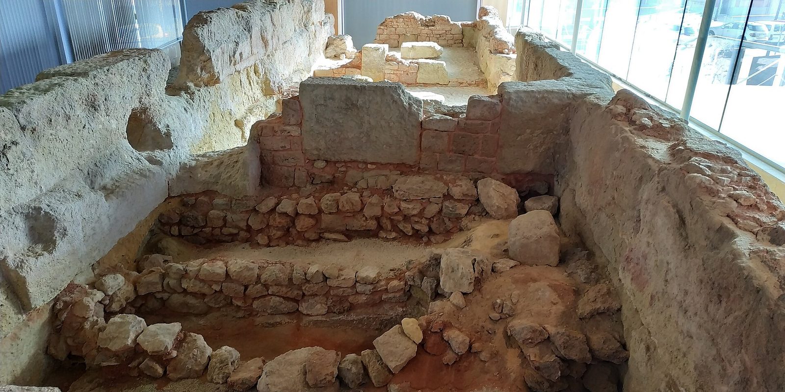 Excavated stone wall footings in a trench under a modern glass-walled shelter.
