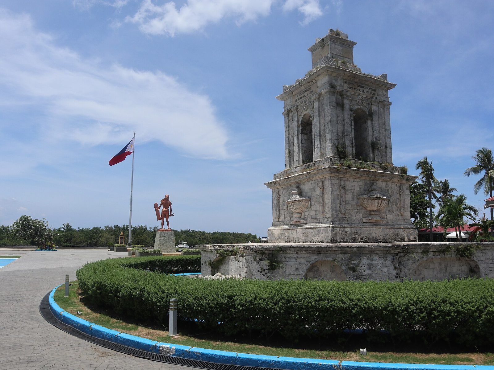 A weathered stone tower monument on a raised base beside a road, and in the distance a bronze statue of a warrior and a flagpole.