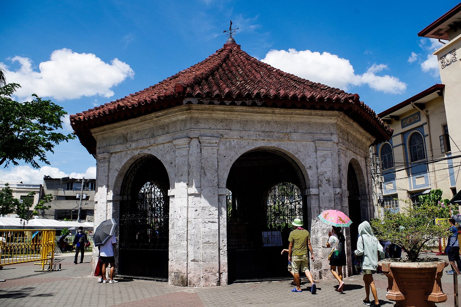 An octagonal stone pavilion with a red tiled roof and arched openings, visitors walking around it under a blue sky.