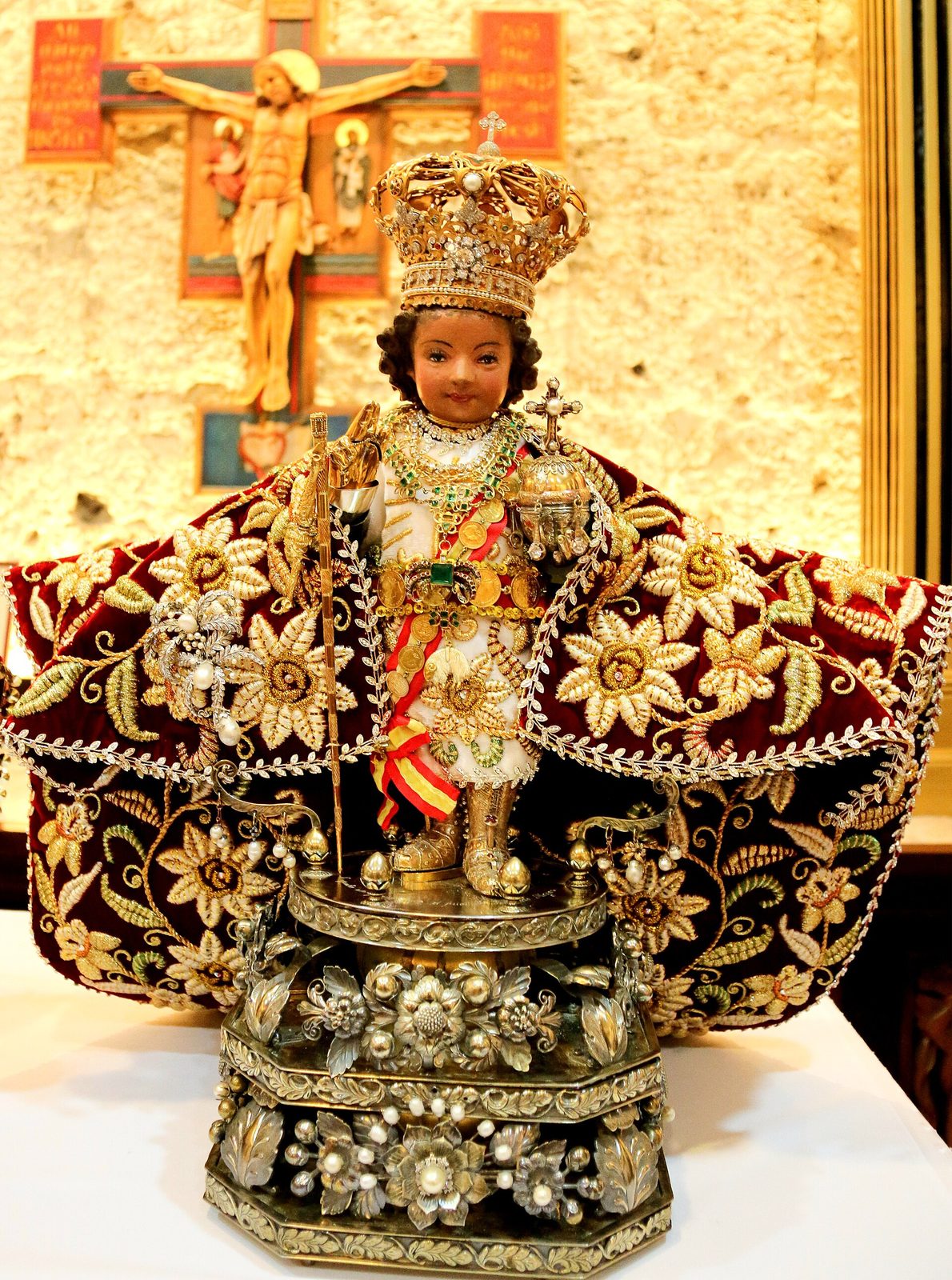 A small carved figure of the Christ child in a jewelled crown and a heavy embroidered red and gold cape, standing on a silver base.