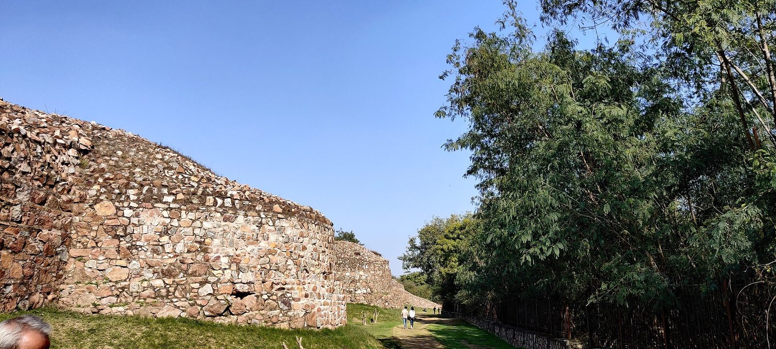 A long rubble-stone fortification wall beside a path and trees.