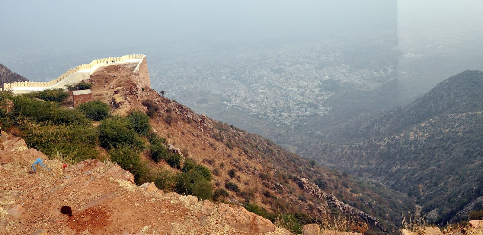 A stone fort wall running along a steep rocky ridge above a city in haze.