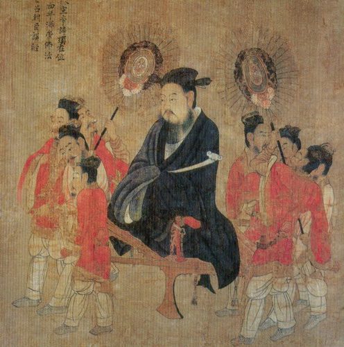 A painted ruler in a dark robe seated on a low platform, surrounded by attendants carrying fans.