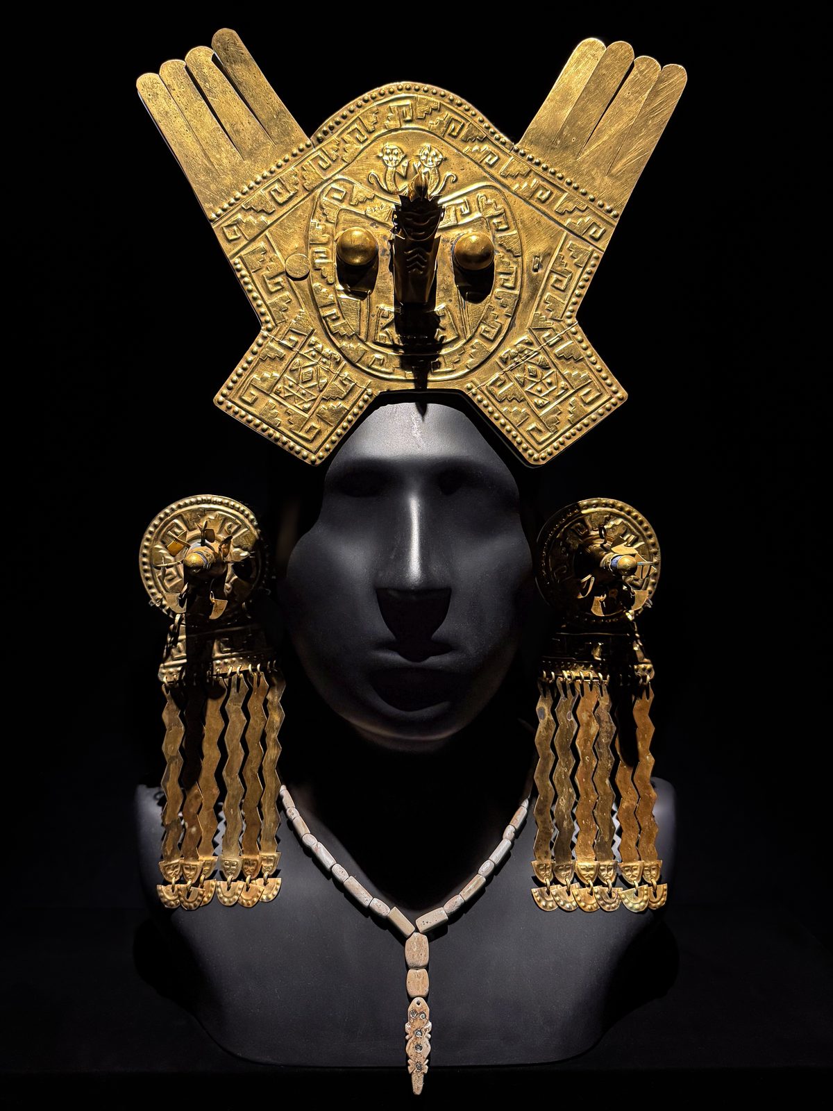 A gold headdress with hand-shaped plumes and long dangling ear ornaments, displayed on a dark mannequin head.