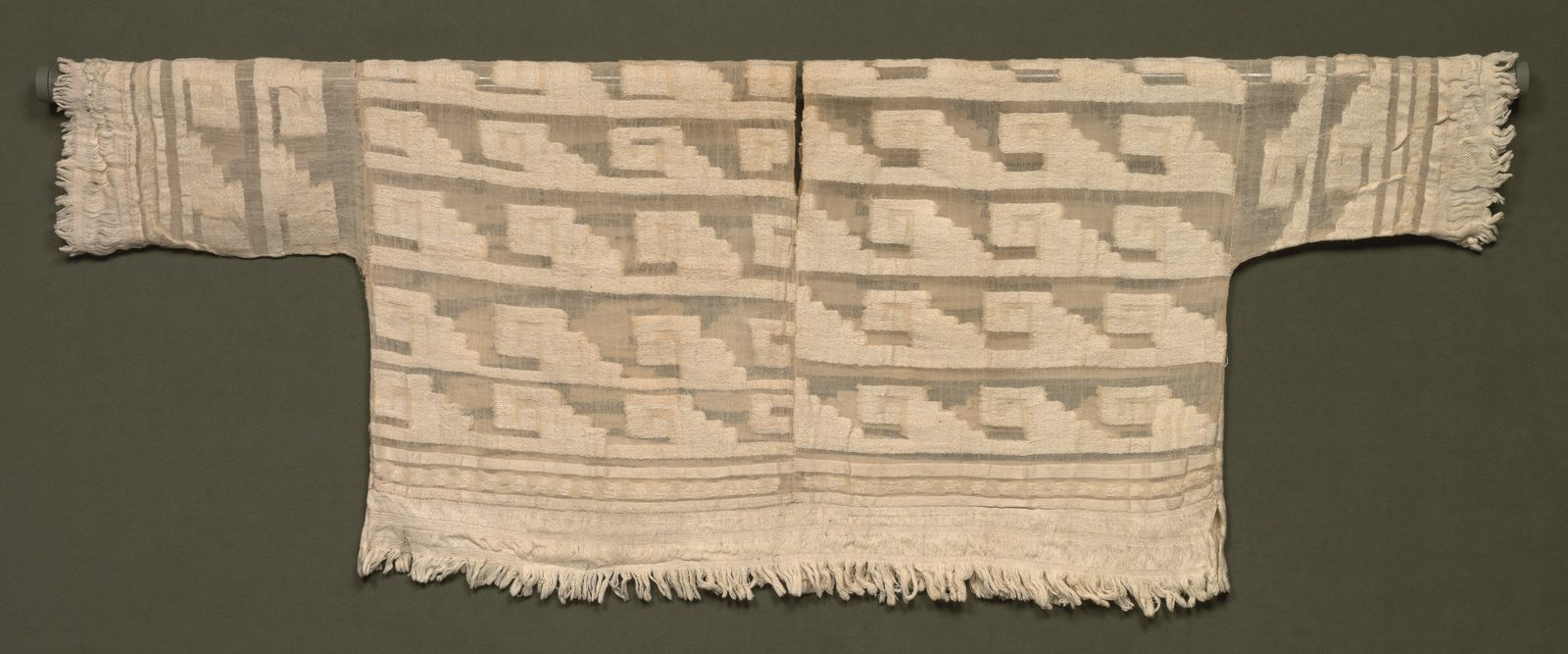 A white cotton tunic woven with rows of stepped and hooked patterns, with a fringe.