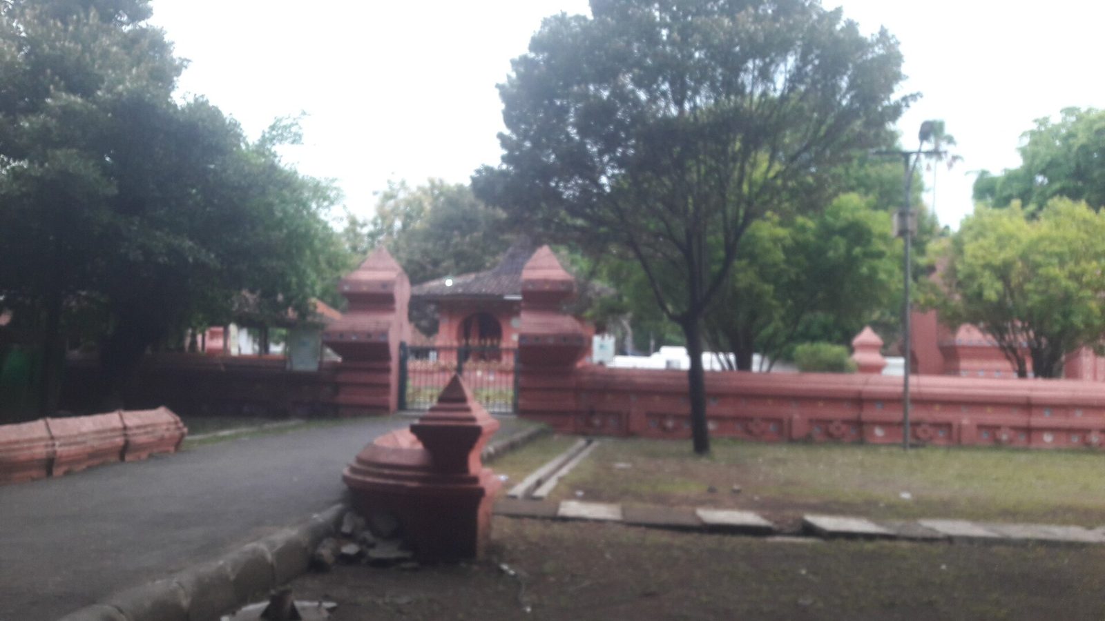 Red-brick walls and a pointed brick gate across a paved yard, with tall trees behind.