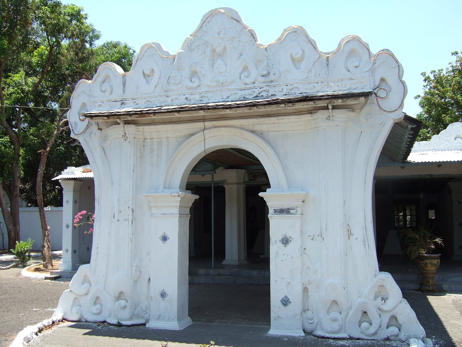 A whitewashed arched gateway with a scalloped crest and carved pilasters standing in a palace yard.