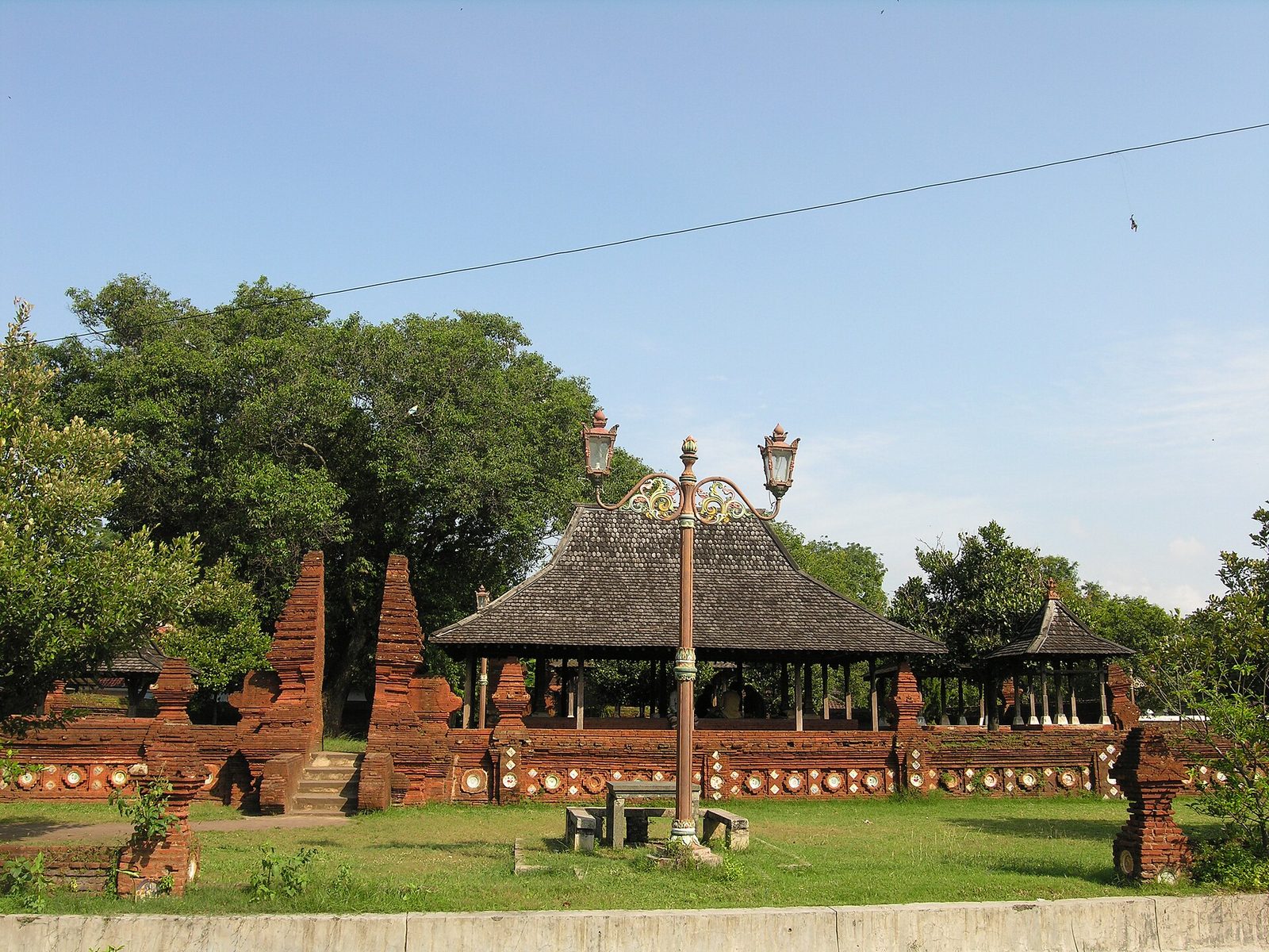 A raised brick terrace enclosed by red-brick split gates and low walls, with a small open pavilion under a thatched roof and trees behind.
