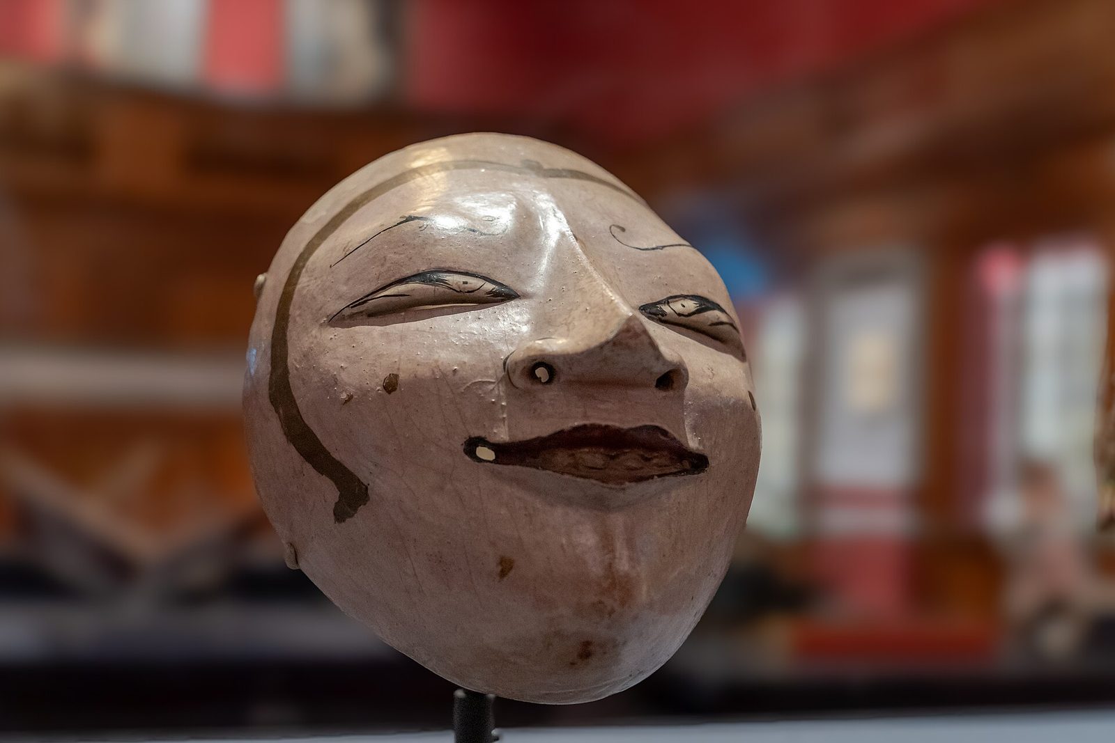 A carved and painted wooden mask with a pale face, narrow eyes and a fine moustache, displayed on a stand.