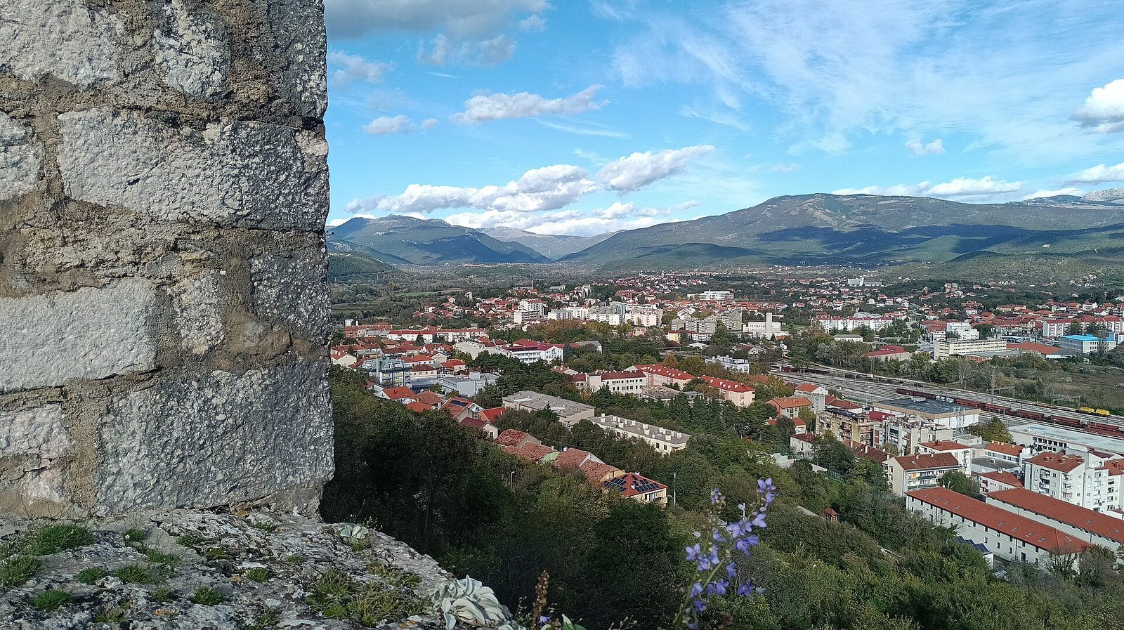 A view from a stone fortress wall over a town in a green valley ringed by mountains.