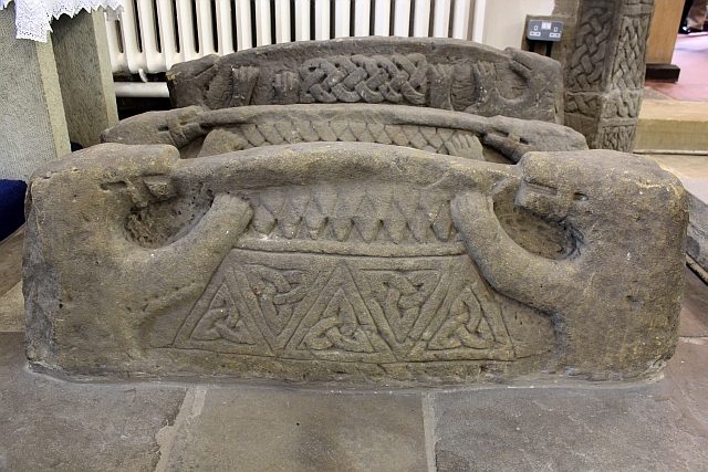 A carved stone grave-cover shaped like a house with a ridged roof, gripped at each end by a beast.