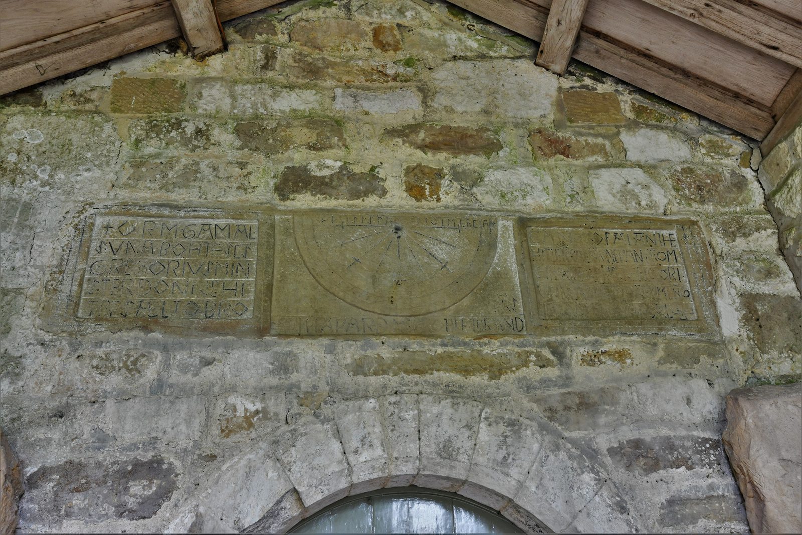 A long stone panel above a church doorway, carved with a sundial between two inscribed panels.