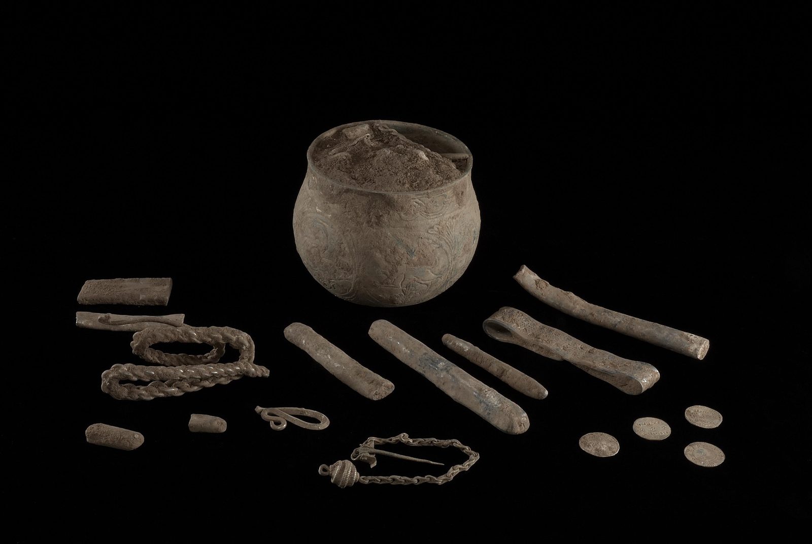A small silver cup surrounded by ingots, arm-rings and coins on a black background.