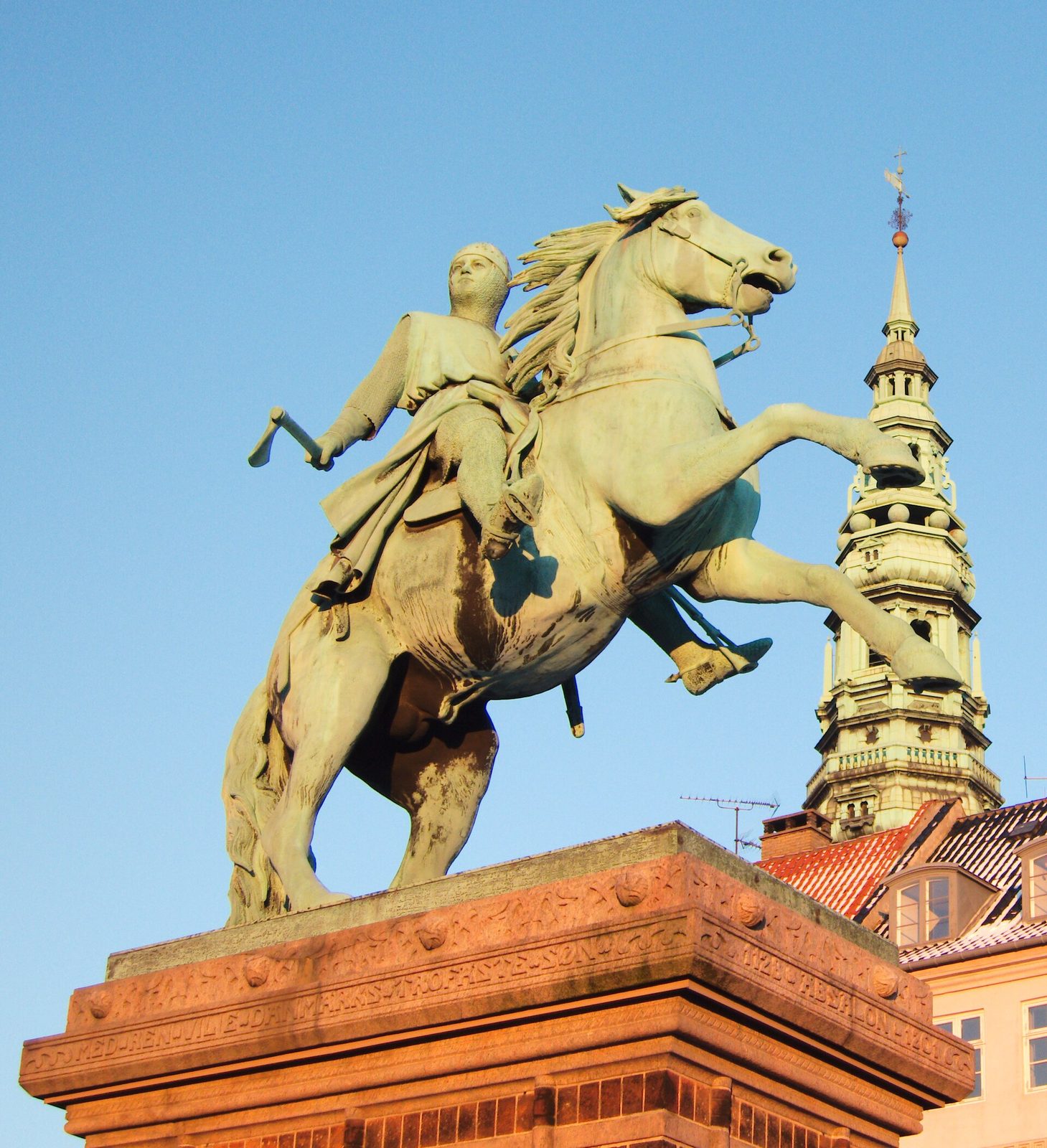 A green bronze equestrian statue of a bishop raising an axe, seen against a blue sky beside a church spire.