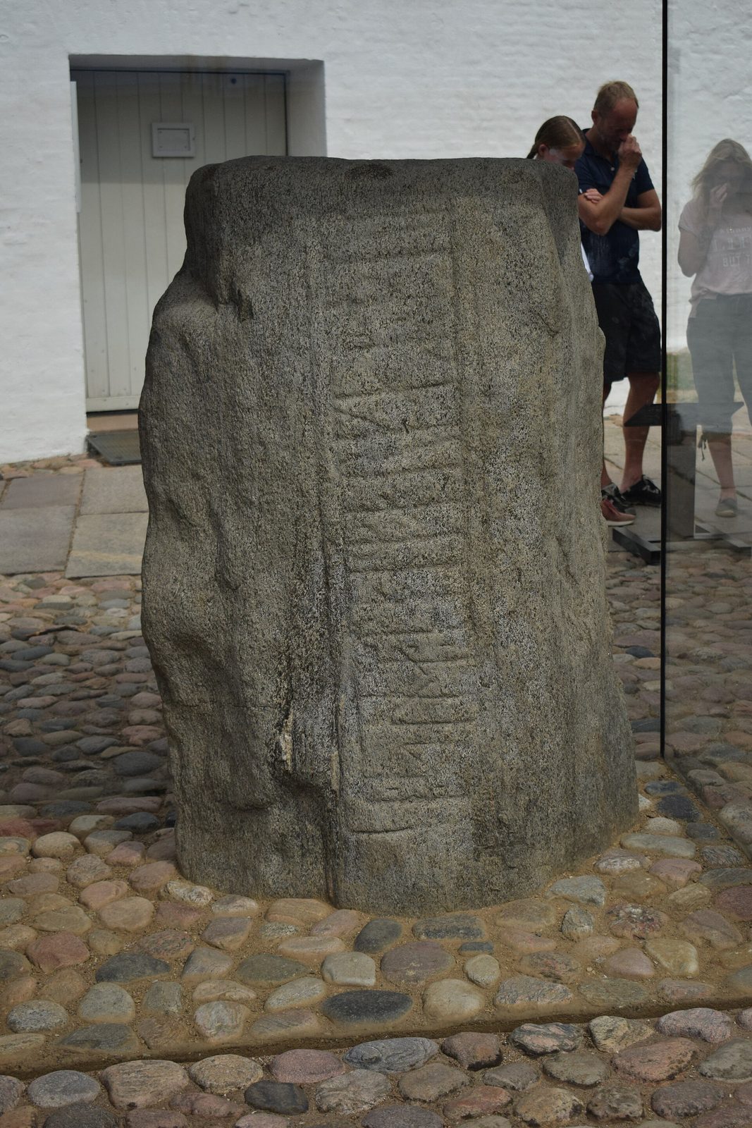 A grey standing stone carved with lines of runes, set on cobbles beside a whitewashed wall.